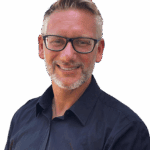 Ein Mann mit kurzen, hellbraunen Haaren, Brille und gestutztem Bart lächelt, während er ein dunkles Button-up-Hemd trägt und bereit ist, Ihnen beim WordPress reparieren zu helfen. Er steht vor einem schlichten weißen Hintergrund.